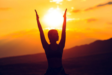 Fototapeta premium Silhouette of young woman practicing yoga or pilates at sunset or sunrise in beautiful mountain location.