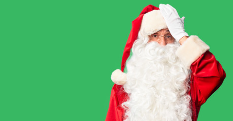 Middle age handsome man wearing Santa Claus costume and beard standing surprised with hand on head...