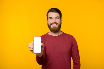 Photo of cheerful guy with beard and wearing red sweater, showing mobile screen at camera over...