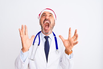 Middle age doctor man wearing christmas hat and stethoscope over isolated white background crazy and mad shouting and yelling with aggressive expression and arms raised. Frustration concept.