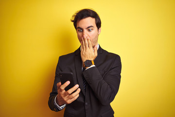 Young handsome businessman using smartphone standing over isolated yellow background cover mouth with hand shocked with shame for mistake, expression of fear, scared in silence, secret concept