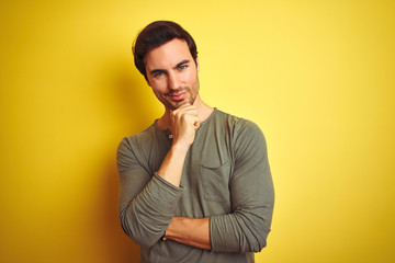 Obraz premium Young handsome man wearing casual t-shirt standing over isolated yellow background looking confident at the camera smiling with crossed arms and hand raised on chin. Thinking positive.
