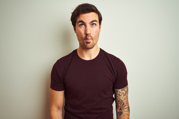Young handsome man with tattoo wearing purple casual t-shirt over isolated white background making fish face with lips, crazy and comical gesture. Funny expression.