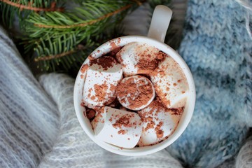 Obraz na płótnie Canvas Christmas hot chocolate or cocoa with marshmallow in white mug on white table with fir branch, cone, knitted blue scarf and gloves. Winter hot drink and christmas decorations. closeup, flatlay