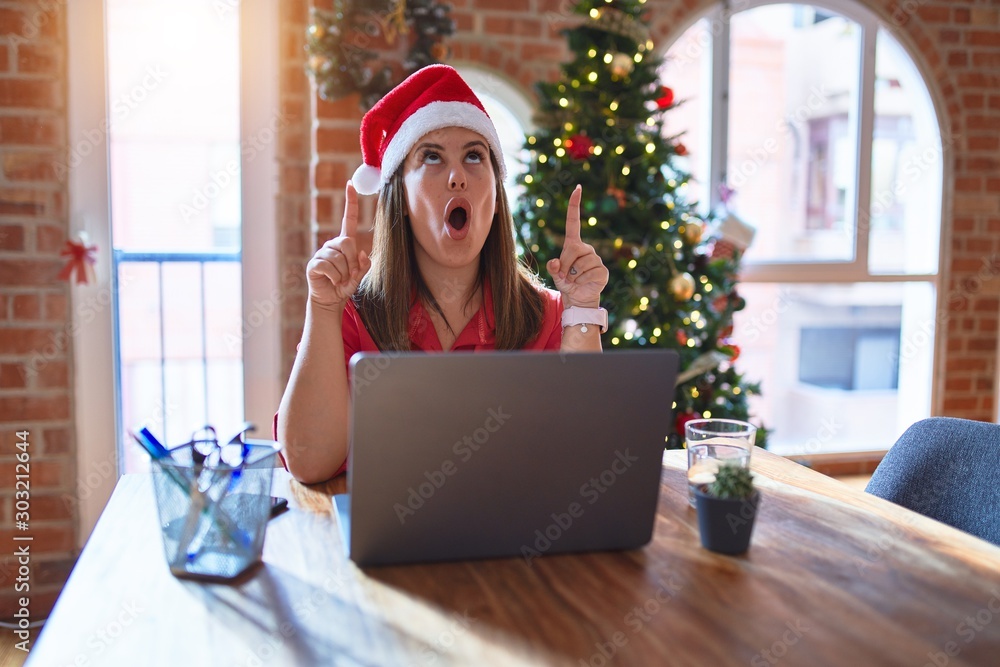 Sticker Beautiful woman sitting at the table working with laptop wearing santa claus hat at christmas amazed and surprised looking up and pointing with fingers and raised arms.