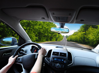 Car dashboard with driver's hands on the steering wheel and rear view mirrors on a road in motion with trees against sky with clouds - 303212627