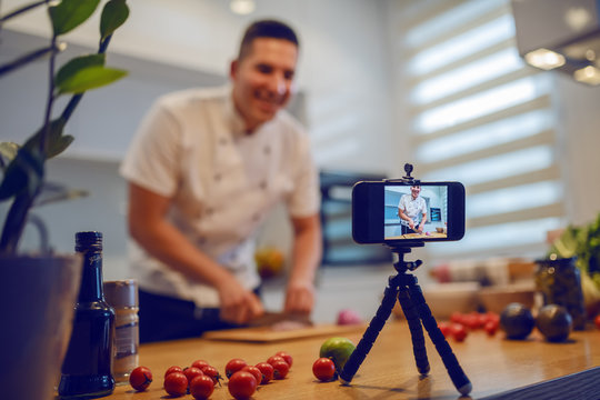 Smiling caucasian chef in uniform standing in kitchen and cutting onion while filming himself for blog. On kitchen counter are vegetables and spices. Selective focus on smart phone.