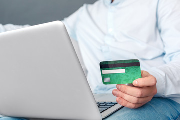 Online Shopping. Young man studio sitting on sofa isolated on grey doing online shopping on laptop paying by debit card for purchase close-up