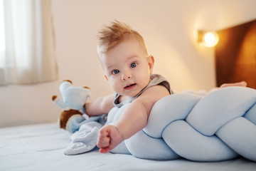 Fototapeta premium Adorable caucasian baby boy with big blue eyes lying on stomach on bed in bedroom and looking curiously at camera.