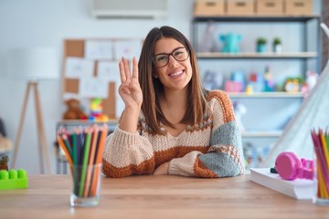 Young beautiful teacher woman wearing sweater and glasses sitting on desk at kindergarten showing and pointing up with fingers number three while smiling confident and happy.