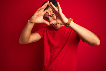 Young brazilian man wearing t-shirt standing over isolated red background Doing heart shape with hand and fingers smiling looking through sign