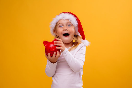 little blonde girl in Santa hat holding piggy Bank on yellow background isolate, space for text