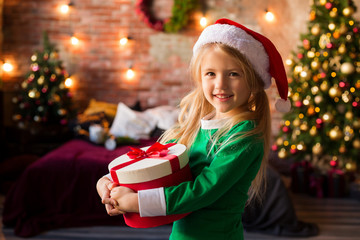 little blonde girl in green pajamas and Santa hat holding a gift box and smiling on the background of a Christmas tree, space for text. merry Christmas!