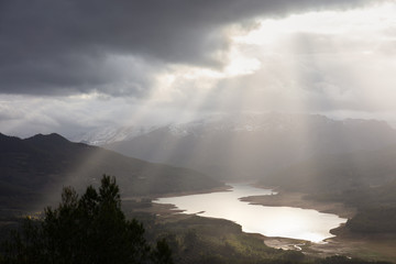 The large reservoir Tranco in the Spanish mountains of Andalusia. The sky is very cloudy and broad sunbeams illuminate the landscape and the lake. It's sunset soon.