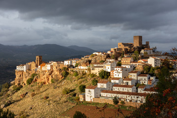 Obraz premium The old historic mountain village of Hornos with white houses in Spanish Andalusia, illuminated by the evening sun in winter. Dark clouds in the sky. There is a church and a castle.
