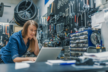 Portrait of beautiful working woman in the bicycle store. Young female mechanic with laptop. Female...