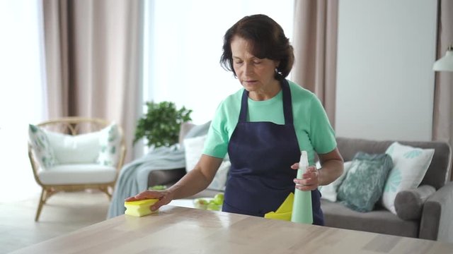 60s Elderly american woman housekeeper wipe table working in home room. 4k Portrait of old female cleaning desk using cleaner, standing in apartment interior with sofa. Employee maintenance cleanness