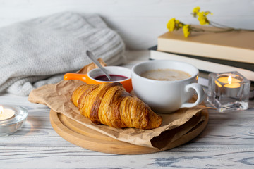 Cozy hygge still life with fresh homemade croissant and coffee on wooden table with candles, books and knitwear