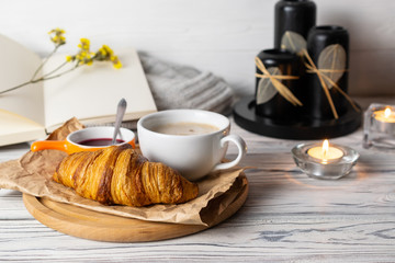 Cozy hygge still life with fresh homemade croissant and coffee on white wooden table with candles, books and knitwear.