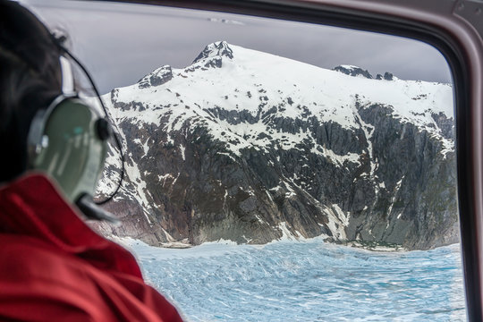 Passenger of the helicopter is looking on the glacier from the window. Amazing snowy mountain peaks. This is excursion in Alaska. Sightseeing tour over the glaciers. Blue icefield.