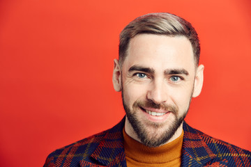 Head and shoulders portrait of smiling bearded man looking at camera while posing against red background, copy space