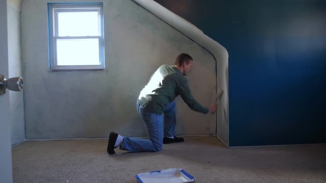 A man in dungarees puts a coat of primer on the walls of a blue room. Wide shot.  	