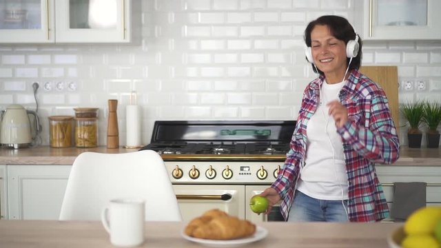 Happy Mature Woman On Headphones Dancing Near Table In Home Kitchen. American Aged Female Holding Apple In Hand And Having Fun, Listening Music Using Headset In Modern Interior. Concept: Healthy