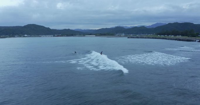 Surfers at Shimosato village coastline waves surfing waves at dusk, Wakayama. Aerial tracking shot of surfer riding wave on surfboard.