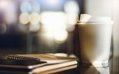 Closeup. Paper cup of hot coffee with notebook and mobile phone on desk. Blurred and bokeh with sunlight background. Concept working and relax with coffee drink