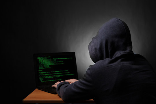 Hacker in hood with laptop initiating cyber attack. View from the back.