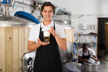 Man brewer in apron standing with glass of beer in brew-house