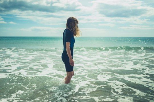 Young woman in wetsuit is standing on the beach with waves crashing in