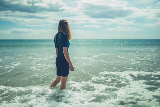 Young woman in wetsuit is standing on the beach with waves crashing in