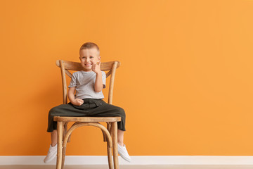 Fototapeta na wymiar Portrait of cute little boy sitting on chair and showing thumb-up gesture against color background