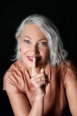 Portrait of stylish mature woman showing silence gesture on dark background