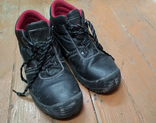 Pair Of Old Black Used Dirty Work Boots on wooden floor. Protective working shoes to protect against accidents at the factory, security tool. Hiking boots, well worn and muddy.