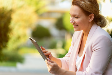 Smiling businesswoman with a tablet in the park. Successful  businesswoman typing text on a  tablet, outdoors. Adult woman communicating with a people uses tablet.