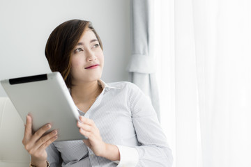 Portrait of a happy Asian woman sitting on sofa using Digital Tablet near window at home.