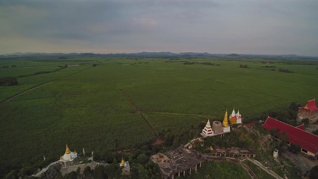 Vdo. Aerial view evening above many Pagodas on top hill around with green sugar cane plantation and cloudy sky background, Wat Nong Kwang Mai (Tham Balami), Photharam, Ratchaburi, Thailand.