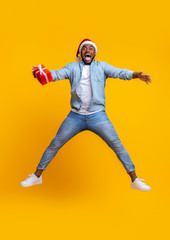 Fototapeta na wymiar Overjoyed afro guy in santa hat jumping with gift box