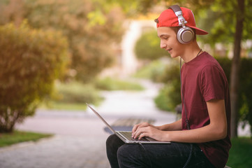 Smiling teenage boy with a laptop in the park. Handsome young man works on a notebook, outdoors....