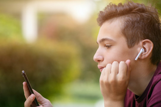 Close-up Portrait Of A Thoughtful Unhappy Teenage Boy With Smartphone, Outdoors.  Sad Teenager With Mobile Phone Looks Away, In The Park.  Pensive Teenager In Casual Clothes With Cell Phone In Park