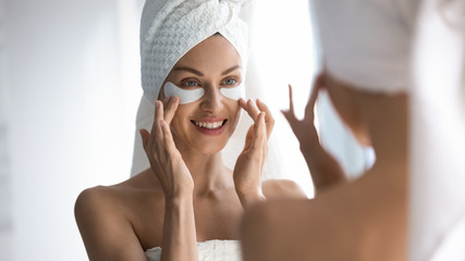 Happy woman towel on head apply patches looking in mirror