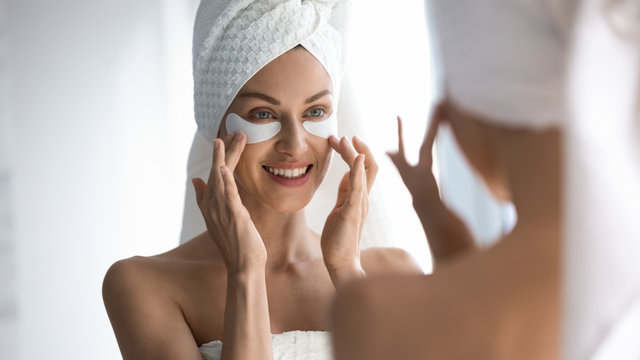 Happy woman towel on head apply patches looking in mirror