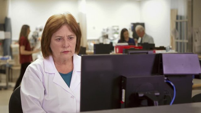 An Mature Female Doctor Using A Modern Computer In A Busy Hospital Setting.