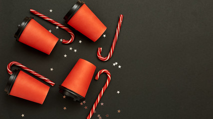 Paper coffee cup with sugar canes on a black background. Christmas background with Xmas candy canes and star confetti. Top view, flat lay, mockup. Copy space for text.