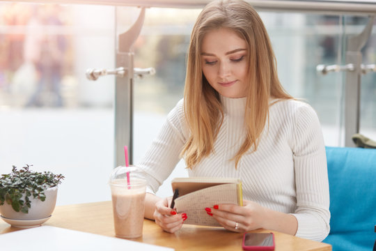 Indoor horizontal picture of sweet good looking young girl holding her notebook, checking notes, looking attentively at pages, wearing white sweater, having cocktail on table. Rest time concept.