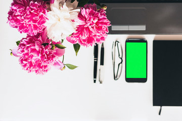 bouquet of pink peonies, laptop, green screen smartphone, pens, glasses and a notebook on a white table, top view.
