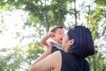 Mom lifting hold infant baby boy in city park against sun light