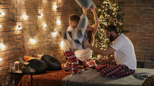 Young bearded dad plays with his children in the bedroom on Christmas evening, a friendly family have fun and fight pillows while sitting on the bed
